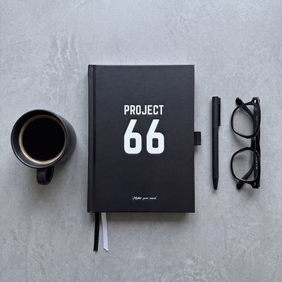 Black notebook with 'Project 66' on a gray surface, accompanied by a coffee cup, pen, and glasses.