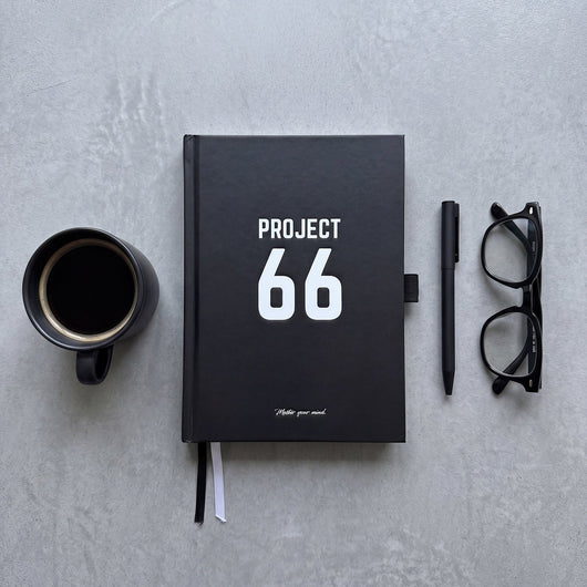 Black notebook with 'Project 66' on a gray surface, accompanied by a coffee cup, pen, and glasses.